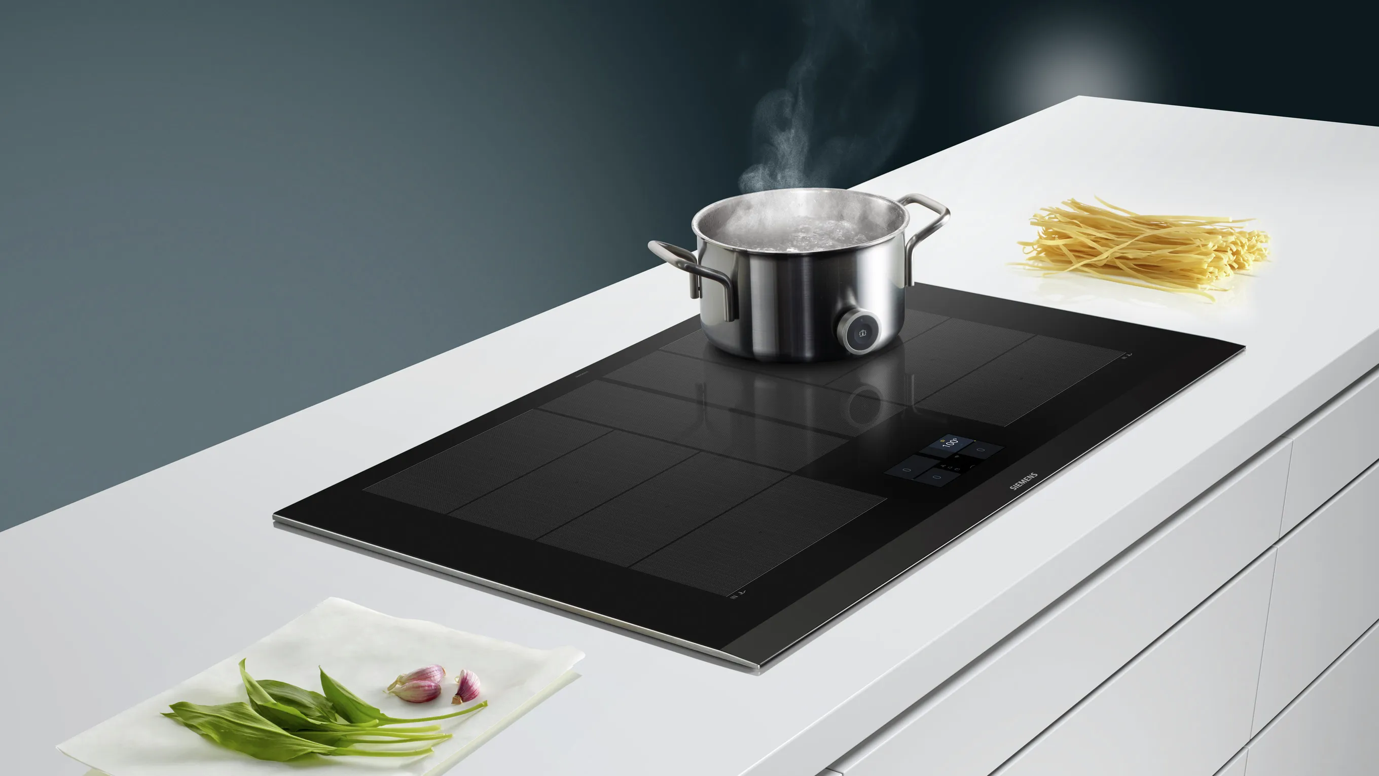 Stainless steel pot on induction cooktop, pasta, and fresh vegetables on white kitchen counter.