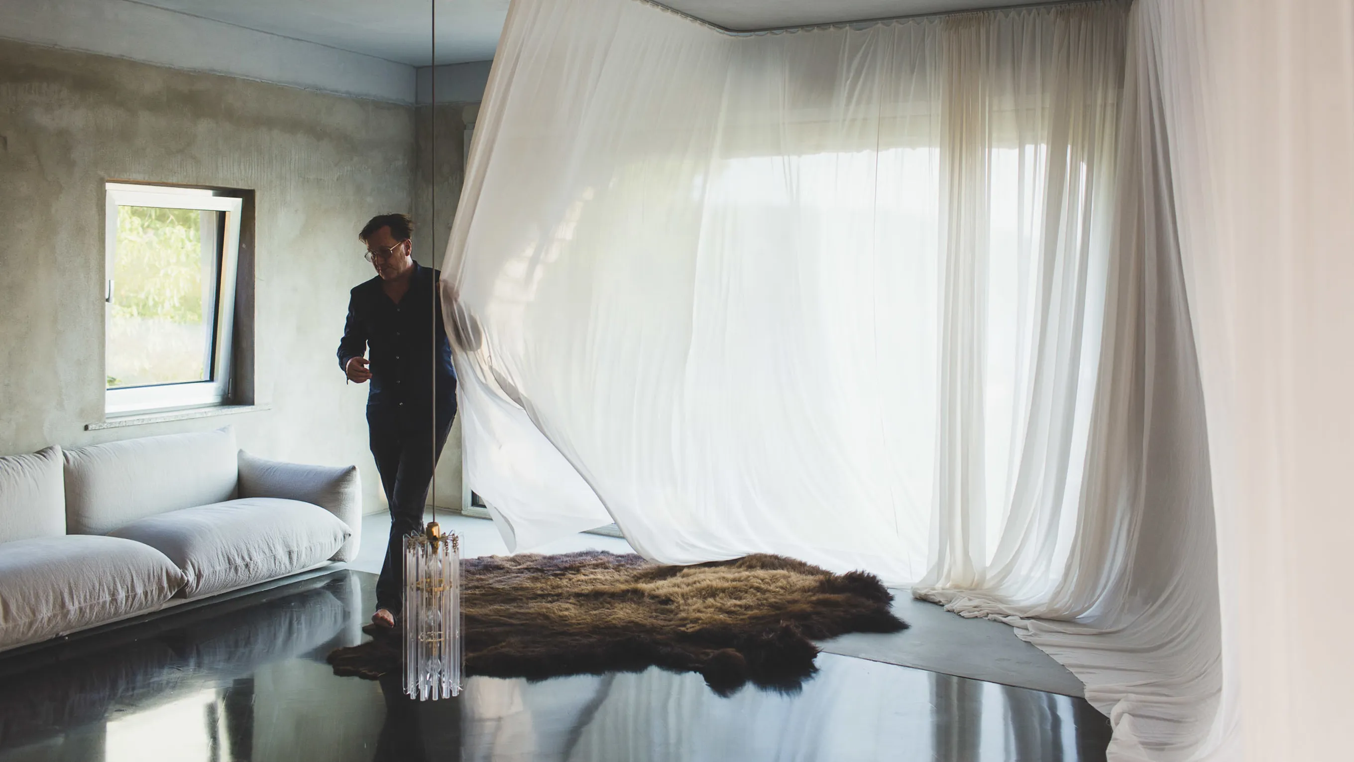 Minimalist bedroom with large window, flowing curtains, and a person standing near a wooden log on the floor.