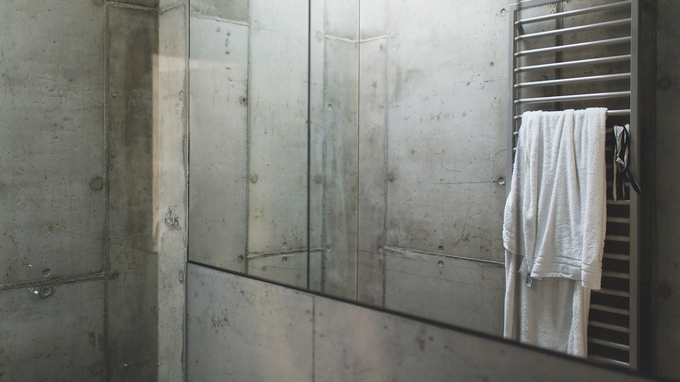 Minimalist bathroom with concrete walls, glass shower enclosure, and a white towel hanging on a metal rack.