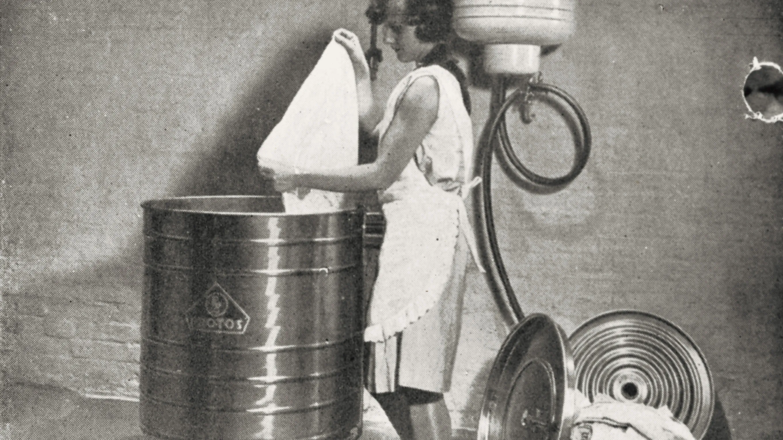 Woman in apron washing dishes in large metal tub, surrounded by various household items.