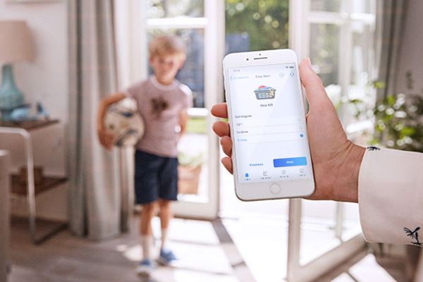 A child standing in the hall with dirty clothes. The mum stops the washing cycle with the Home Connect app.