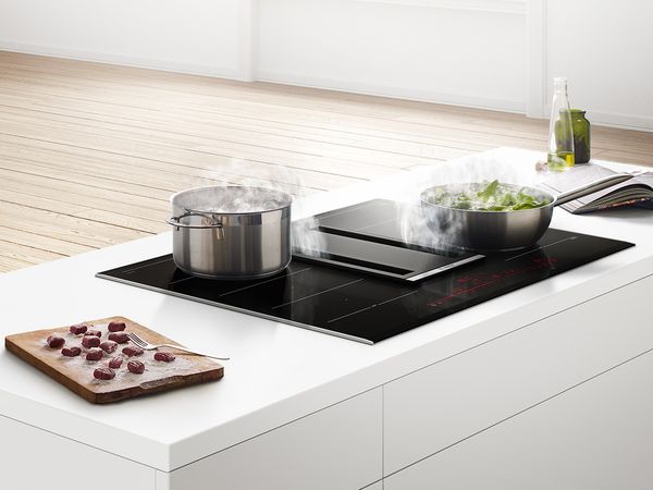 Table de cuisson et hotte intégrée en position centrale dans une cuisine; des aliments cuisent dans une poêle et de l'eau boue dans une casserole.