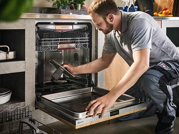 Un technicien est agenouillé devant un lave-vaisselle ouvert et retire un panier, dans une cuisine moderne avec des fruits sur le comptoir.