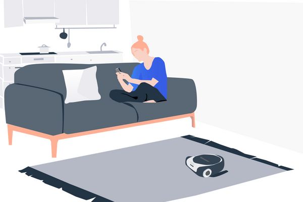 一位女士正在沙發上看著她的智慧型手機，同時吸塵機器人則在打掃地毯。