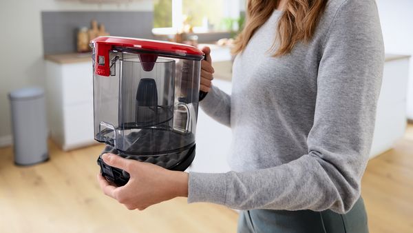 Eine Frau tr&auml;gt den Beh&auml;lter eines beutellosen Staubsaugers von Bosch in ihrer Hand.