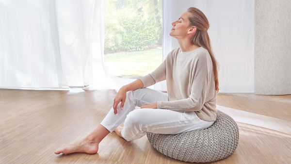 Eine blonde Frau barfu&szlig; in Wellness-Kleidung auf einem grauen Strick-Sitzkissen in einem Raum mit Parkettboden und wei&szlig;en Gardinen.