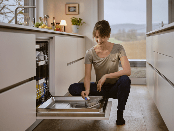 Hölgy térdel a mosogatógép előtt a konyhában, amint egy mosogatógép-tablettát tesz be a mosogatógépbe.