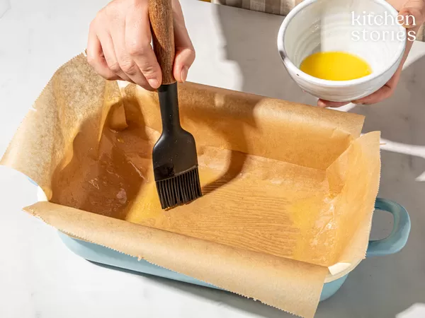 Hands brushing melted butter into a parchment-lined loaf pan with a silicone pastry brush, a small bowl of melted butter held in the other hand; "kitchen stories" logo in the top right.