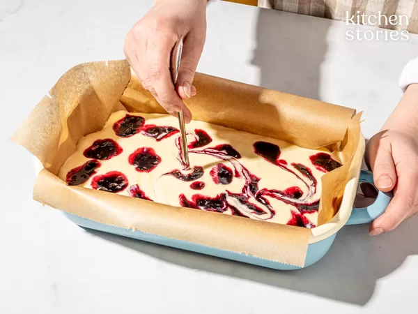 Hands holding a parchment-lined blue loaf pan while one hand swirls dark red jam into pale batter with a skewer, creating marbled swirls; "kitchen stories" logo visible in the top right.