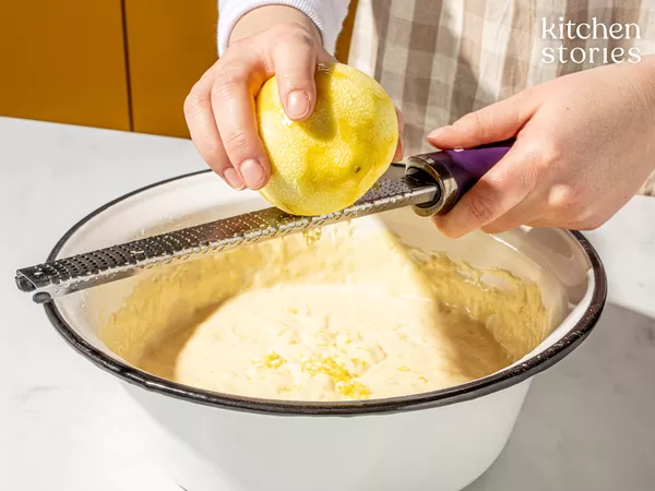 Two hands grating lemon zest with a microplane over a white enamel bowl of batter, bright yellow zest falling into the mixture; "kitchen stories" logo in the top right.