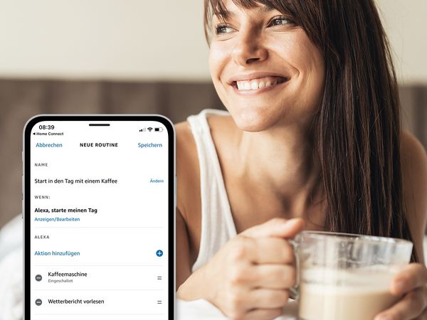 Eine Frau liegt mit einem Kaffeegetränk in der Hand lächelnd im Bett. Smartphone mit Display der Routine in der Alexa App im Vordergrund.