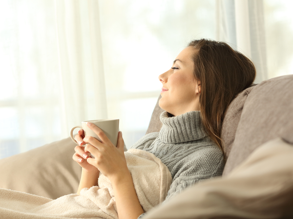En dame ligger smilende i sengen med en kaffedrikk i hånden. Smarttelefon med skjermbilde av en rutine i Alexa-appen i forgrunnen.