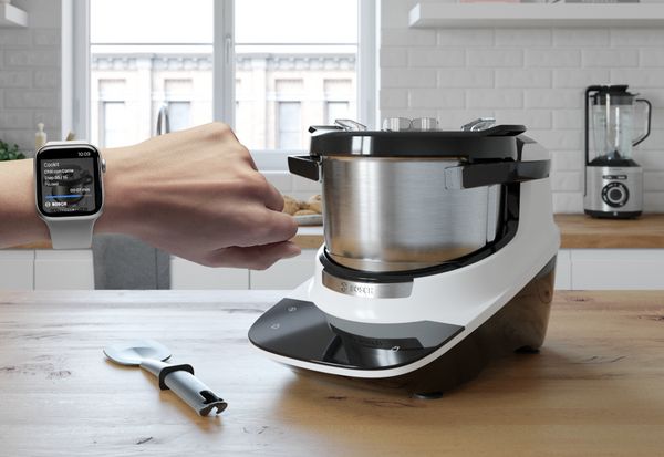 Ein Arm mit einer Apple Watch am Handgelenk wird in das Bild gestreckt, während im Hintergrund ein Cookit in einer Küche zu sehen ist.