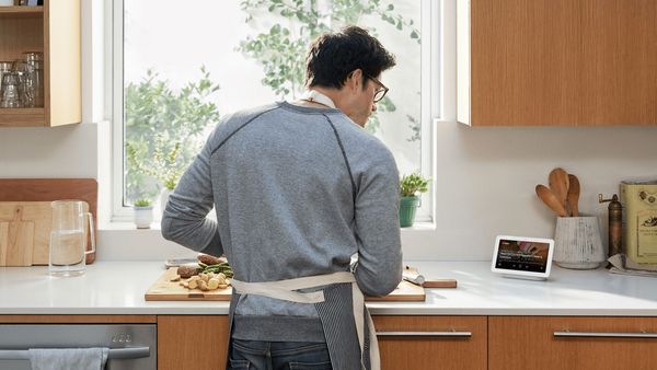 A man cutting assorted vegetables at a chopping board, standing next to a Hey Google device at the countertop