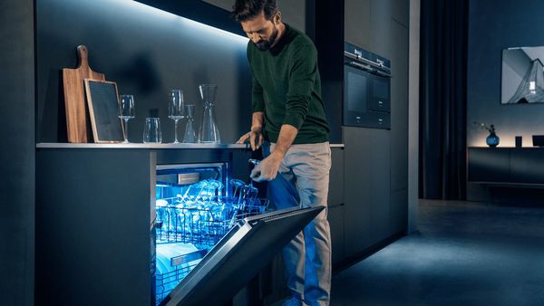 A man in a futuristic-looking apartament furnished in dark blue taking out glasses out of a dishwasher.