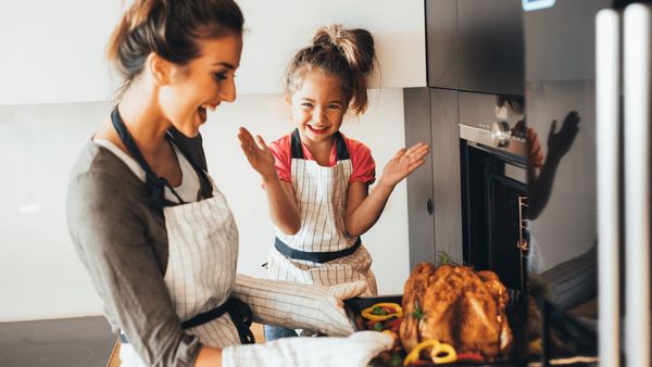 A woman taking out a roast chicken out of the oven while her daughter is clapping.