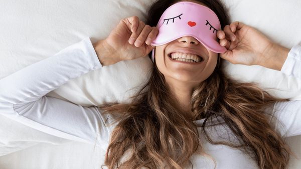 A smiling woman wearing a pink eye sleep mask lying in bed.