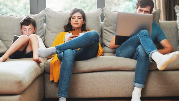 A teenage boy, a woman and a man sitting on a couch – all looking at screens of their respective devices.