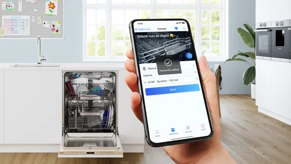 A hand holds up a smartphone showing the silent dishwashing cycle in the Bosch Home Connect app. A loaded dishwasher is visble in the background.