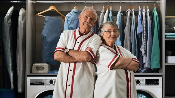 Two spry pensioners in matching white tops stand back to back in an elegant laundry room.