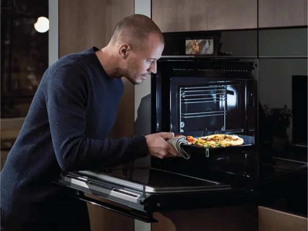 Man in dark sweater removing pizza from oven in kitchen.
