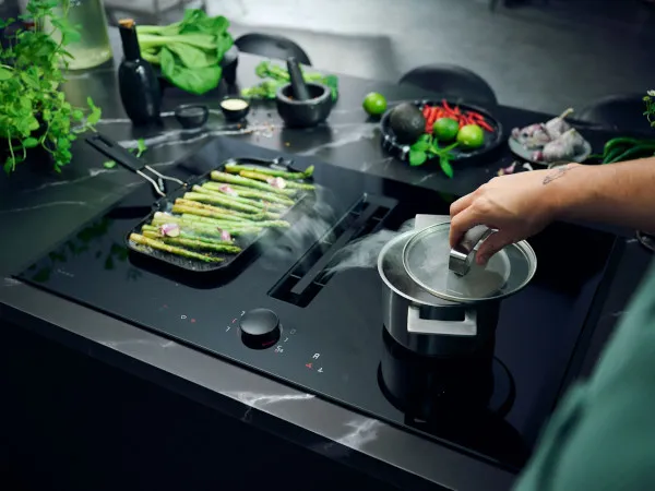 Kochen von Zutaten und Küchengeräten auf einer schwarzen Arbeitsplatte, darunter Gemüse, Kräuter und eine Kaffeemaschine.
