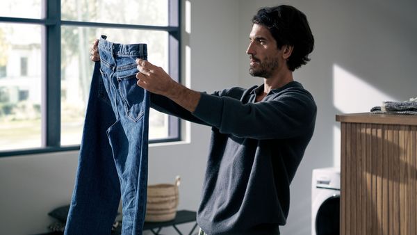Mann steht in der Waschküche und hält eine Jeans in der Hand.