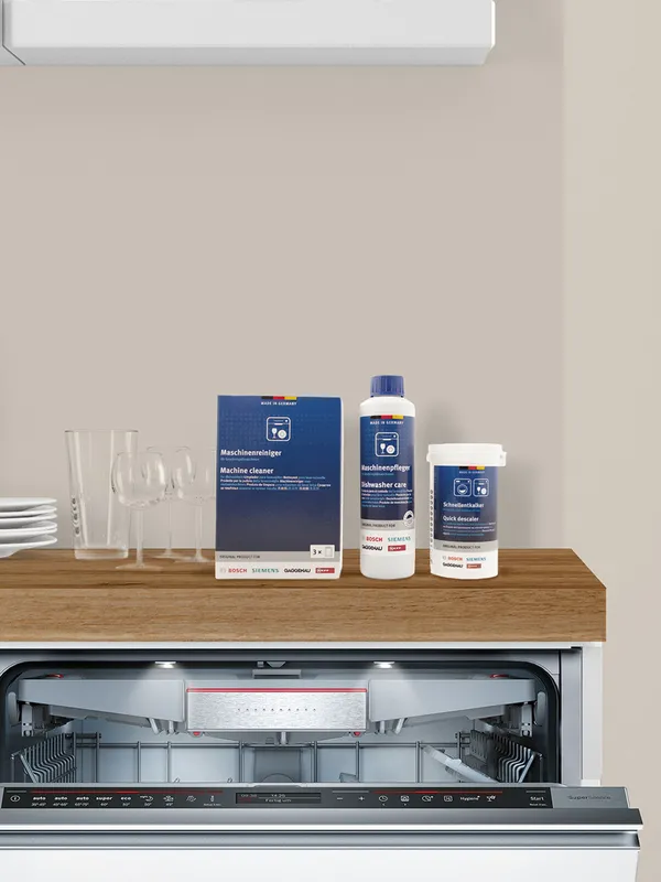 Dishwasher with cleaning supplies on wooden counter, including bottles and glasses.