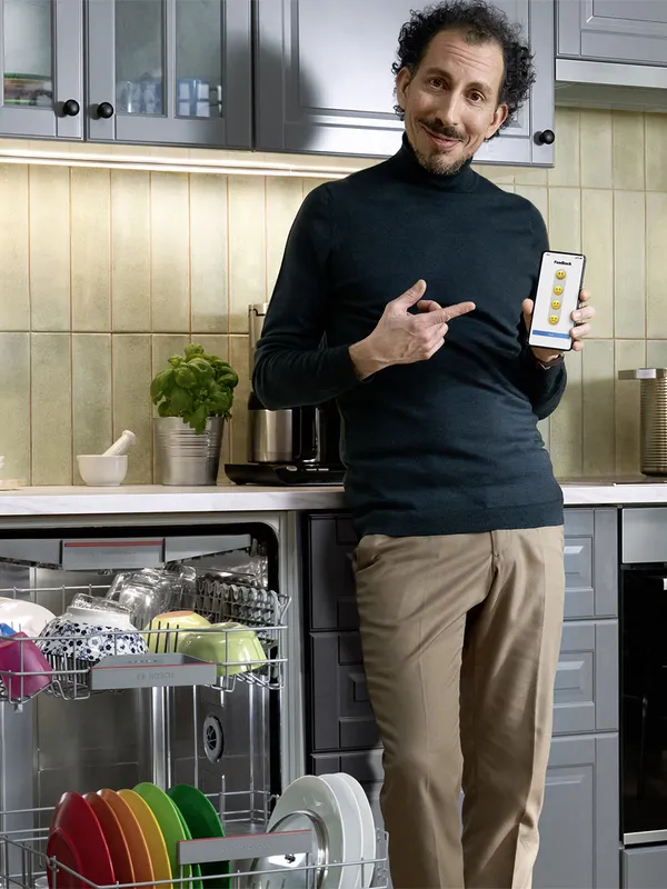 A person in a kitchen is standing next to a dishwasher full of colourful dishes, pointing to a smartphone in her hand.