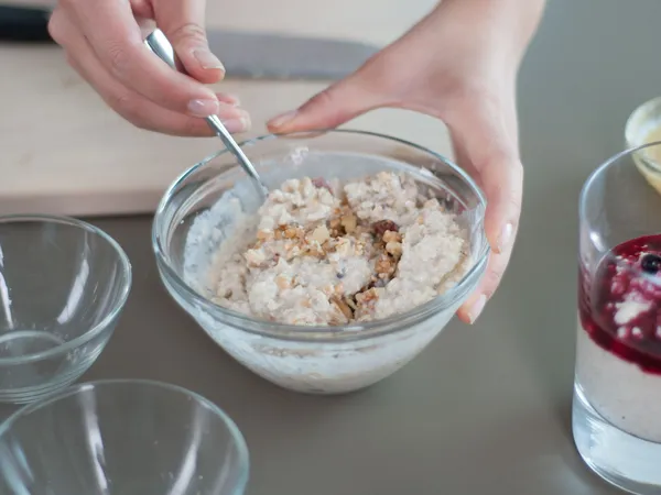 Hands stirring a glass bowl of oatmeal with chopped walnuts on a countertop; a nearby glass holds oatmeal topped with berry compote.