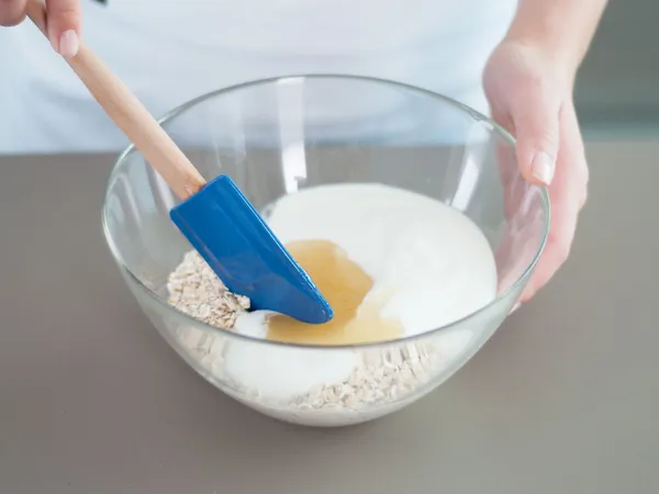 Close-up of hands holding a clear glass mixing bowl with oats, yogurt, and a drizzle of honey, a blue silicone spatula resting inside