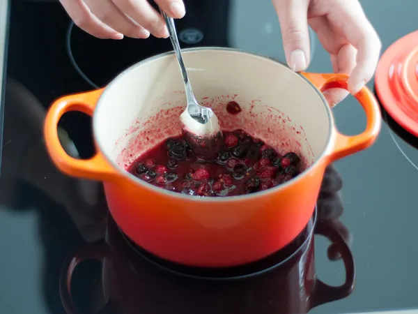 Close-up overhead view of two hands mashing mixed berries with a spoon inside an orange enameled pot on a black stovetop; the pot lid sits nearby