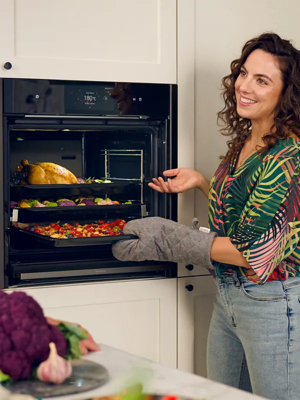 Woman checking on food in oven, smiling, kitchen setting with vegetables on counter.