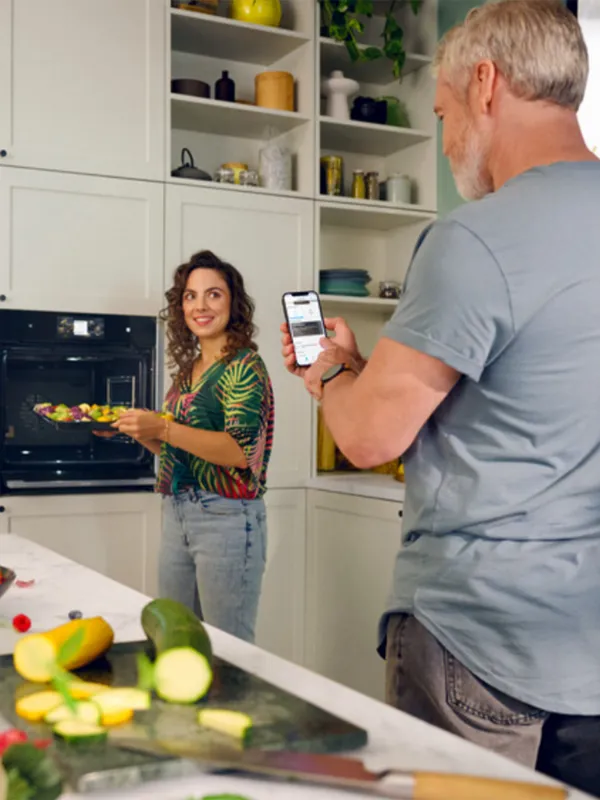 Couple in kitchen, woman cooking, man using smartphone, vegetables on counter.