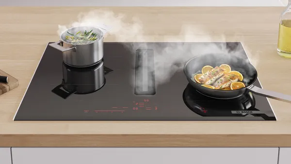 Modern black induction cooktop with pot boiling vegetables and frying pan cooking fish with lemon slices, steam rising.