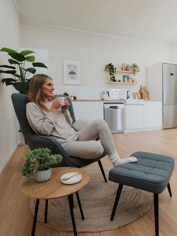 Un espace de vie confortable avec une femme assise dans une chaise moderne, tenant une tasse, avec une table basse, une plante et la cuisine en arrière-plan.