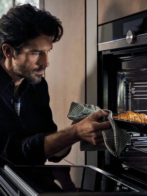 Person using a cloth to take freshly baked croissants out of a modern kitchen oven.