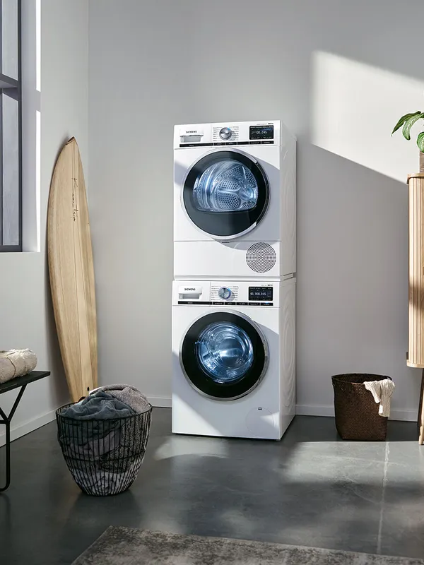 Stacked white front‑loading washer and dryer in a bright laundry corner with a surfboard, baskets, and sunlight.