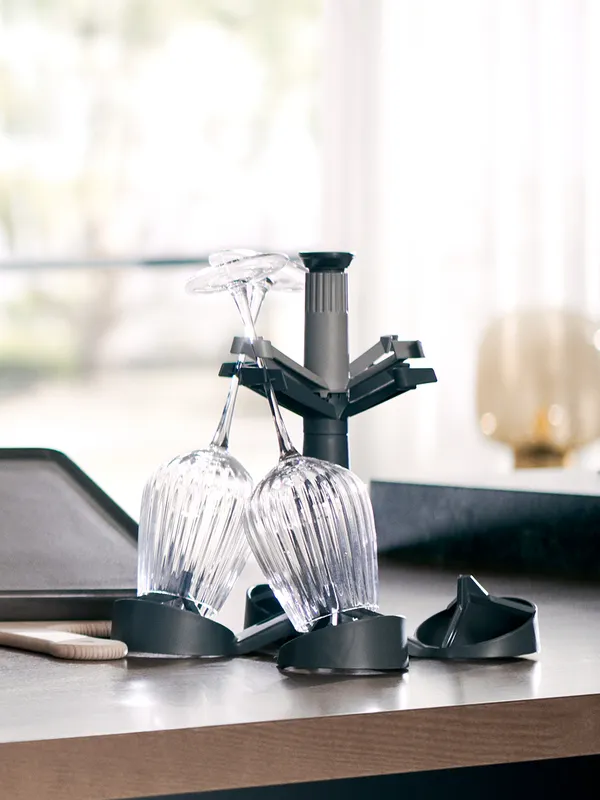 Two inverted crystal wine glasses resting on a black plastic drying rack on a kitchen countertop with a bright, blurred window in the background.