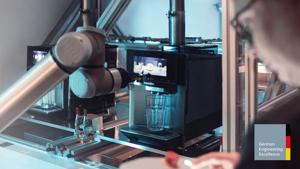 A robotic arm interacts with a Siemens coffee machine in a testing environment, with a person observing and a "German Engineering Excellence" badge.