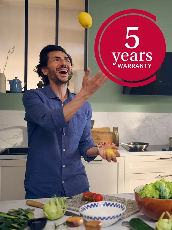 Man in kitchen holding 5-year warranty sign, smiling, surrounded by fresh produce.