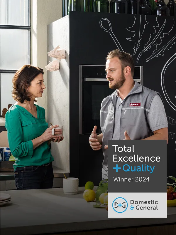 Two people conversing in front of a "Total Excellence in Quality" award display.