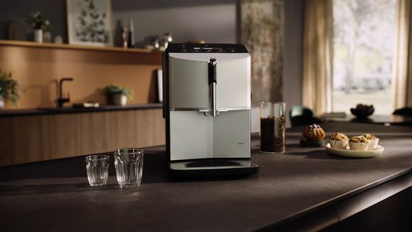 Elegantní kávovar na tmavé desce, vedle něj dvě sklenice, sklenice zrnkové kávy a talíř s muffiny v útulném kuchyňském prostředí.