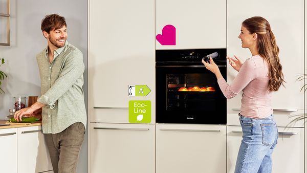 Ein Mann und eine Frau in einer modernen Küche, der Mann schneidet Gemüse auf einem Schneidebrett, während die Frau einen Herd überwacht. Der Herd hat ein Eco-Line Label mit Energiesparkennzeichnung A.