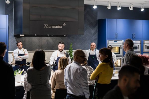 A group of people watch a culinary presentation in a modern kitchen setting, with chefs demonstrating cooking techniques.