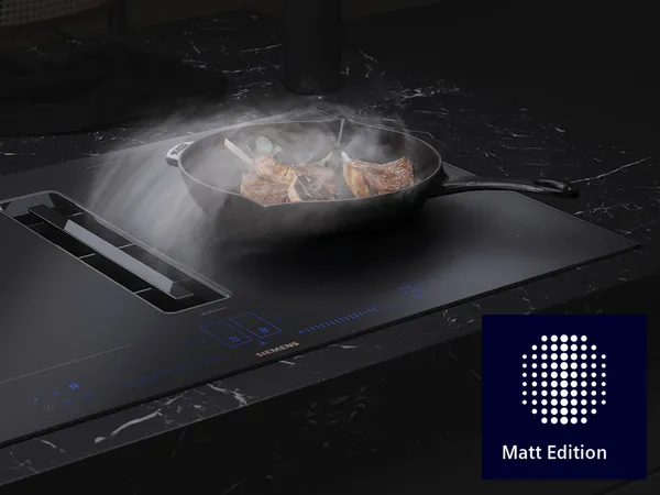 Ein elegantes schwarzes Induktionskochfeld mit einer gusseisernen Pfanne, in der Fleisch angebraten wird, umgeben von Dampf, mit der Marke „Matt Edition“.