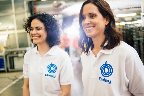 Dos mujeres con camisetas tipo polo blancas con logotipos azules de "Balay" están de pie en un entorno industrial luminoso, conversando entre ellas.