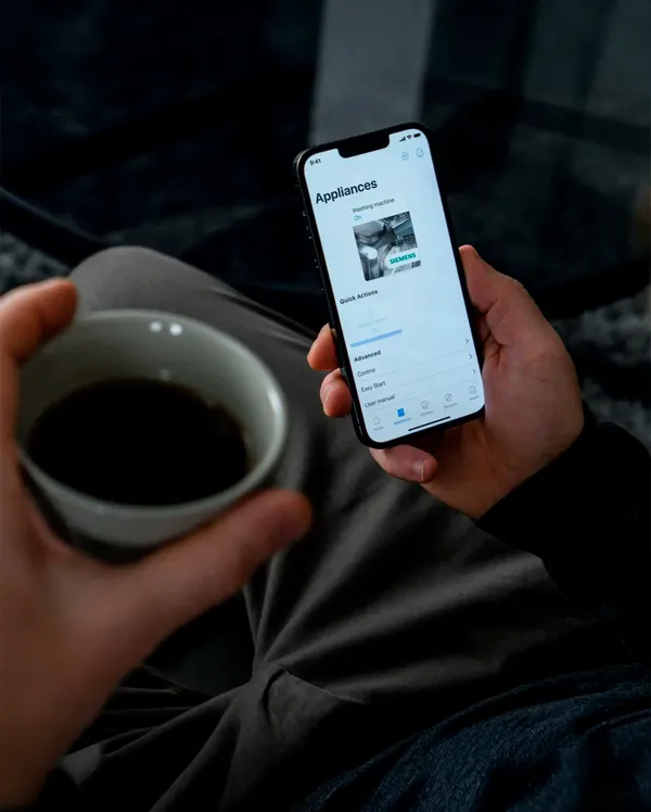 A person holds a coffee cup in one hand while using a smartphone to manage smart home appliances.