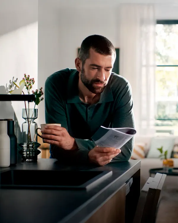 Ένα άτομο σε μια ζεστή κουζίνα στηρίζεται στον πάγκο, κρατώντας μια κούπα και ένα εγχειρίδιο.
