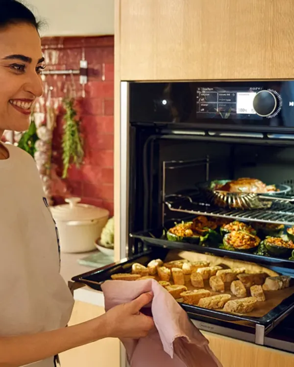 Une femme souriante fait cuire divers plats dans un four moderne et élégant dont la porte est ouverte.